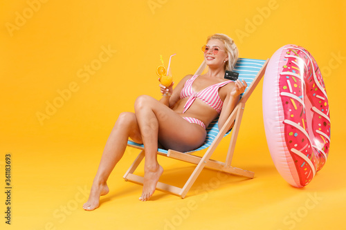 Smiling young woman girl in pink striped swimsuit glasses sit on deck chair isolated on yellow background. People summer vacation rest lifestyle concept. Hold glass with cocktail  credit bank card.