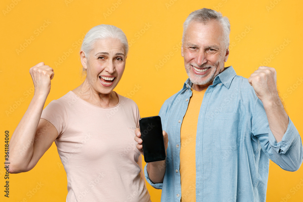 Happy elderly gray-haired couple woman man in casual clothes isolated on yellow background studio portrait. People lifestyle concept. Hold mobile phone with blank empty screen doing winner gesture. Happy elderly gray-haired couple woman man in casual clothes isolated on yellow background studio portrait. People lifestyle concept. Hold mobile phone with blank empty screen doing winner gesture.
