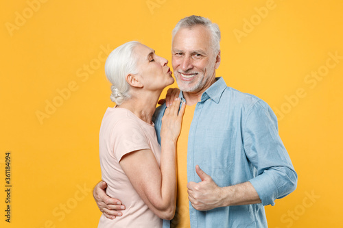 Smiling elderly gray-haired couple woman man in casual clothes posing isolated on yellow background studio portrait. People lifestyle concept. Mock up copy space. Hugging kissing showing thumb up.