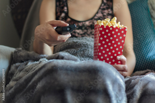 Woman wrapped in warm blanket holding tv remote control and holding box of popcorns