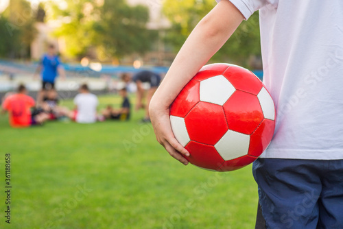 A boy stands on the football field of the stadium and holds a soccer ball against the background of his team or the opposing team. Training or competition concept