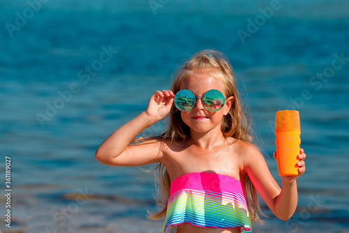 Beautiful little girl in a swimsuit and sunglasses stands by the sea and holds a sunscreen in her hands.