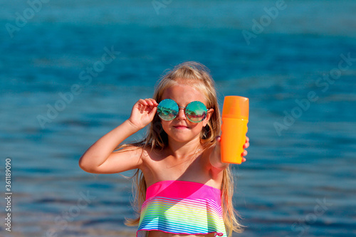 Beautiful little girl in a swimsuit and sunglasses stands by the sea and holds a sunscreen in her hands.