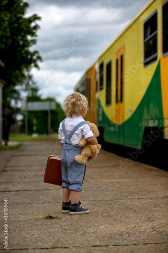 Sweet toddler child with teddy bear, book and vintage suitcase waiting for the train on a train station