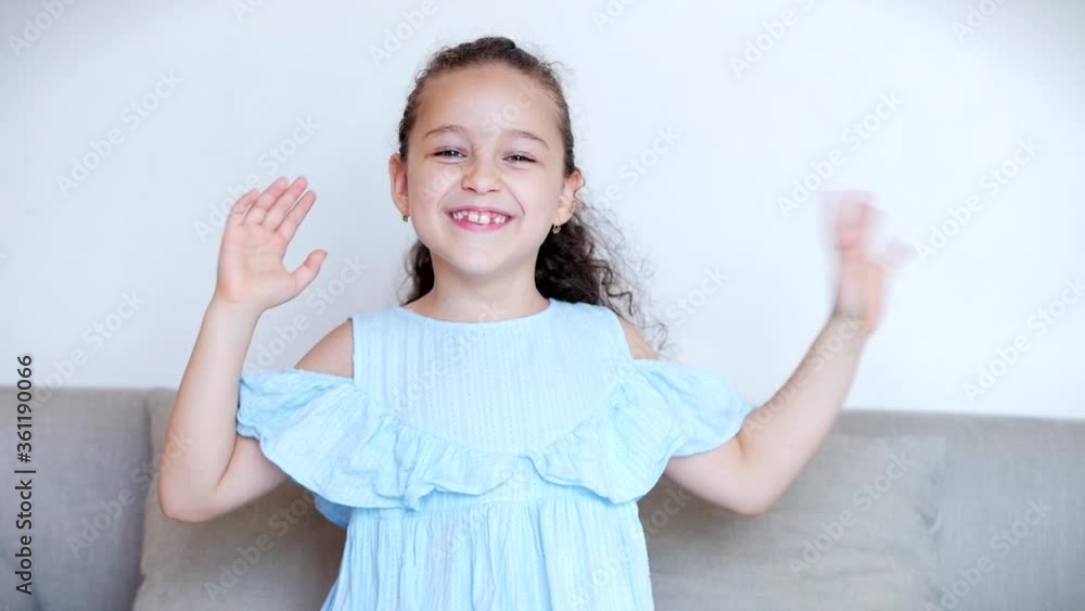 Portrait of a funny little girl smiling. Chill, looking at the camera, waving hands. Hello as a blogger sitting on a sofa at home, cute baby child with a cute face sitting on sofshaw thumb up class Portrait of a funny little girl smiling. Chill, looking at the camera, waving hands. Hello as a blogger sitting on a sofa at home, cute baby child with a cute face sitting on sofshaw thumb up class