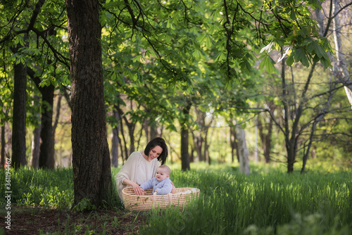 The concept of motherhood and child care. A young mother plays with her little son, who is sitting in a wicker straw cradle. Walk in the park without a stroller sitting on the grass under a chestnut © farmuty
