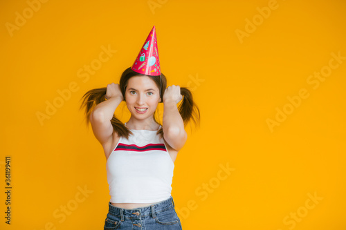 Young woman in a celebratory cap fooling around at a party on the yellow background