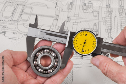 Measurement of steel ball bearing by precise analog caliper above a technical drawing. Metallic measuring tool with round yellow dial in engineer hands. Drafting of combustion engine. Quality control.