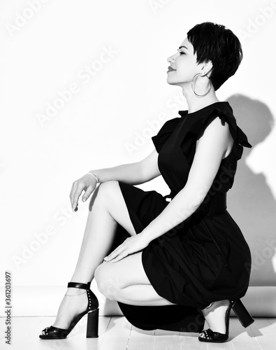 Young short haired brunette woman in elegant black dress, shoes and earrings sitting on floor and smiling