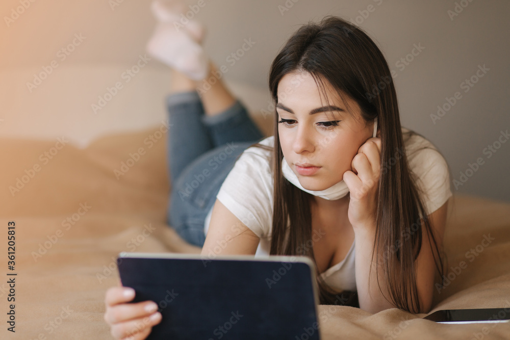 Young woman in mask use tablet on the bed. Female watch online video during quarantine. Student Study at home Young woman in mask use tablet on the bed. Female watch online video during quarantine. Student Study at home