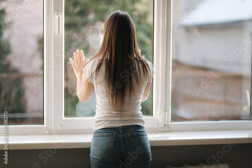 Back view of young woman in medical mask stay by the window in quarantine. Woman looking through the window. Covid-19