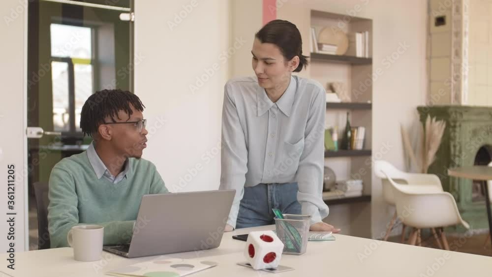 Medium shot of young African man wearing eyeglasses sitting at desk at laptop and communicating with young Asian woman standing nearby Medium shot of young African man wearing eyeglasses sitting at desk at laptop and communicating with young Asian woman standing nearby