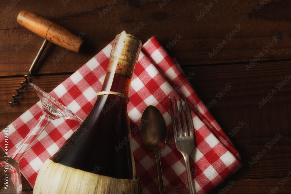 Flat Lay Wine Still Life: A basket bottle of Chianti wine on a red and white checked napkin with corkscrew, fork and spoon with warm sie light. - obrazy, fototapety, plakaty 