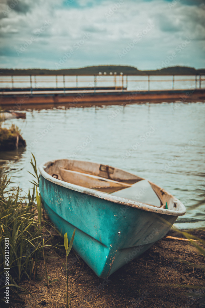 A turquoise boat lies on its side on the shore of a pond next to tall grass. Blurred lake background