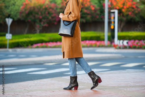 Fashionable young woman in black cowboy boots, blue jeans, beige coat, white t-shirt and crocodile print handbag in hand on the city street. Street style.