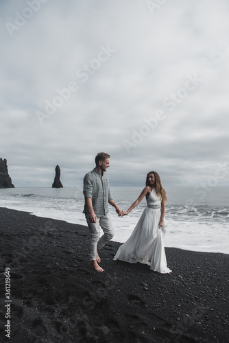 Destination Iceland wedding. A wedding couple is walking along the black beach of Vic. Sandy beach with black sand on the shores of the Atlantic Ocean. Huge foamy waves. The groom hugs the bride.