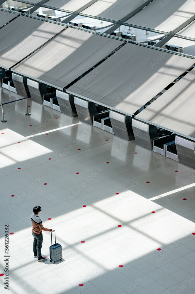 Obraz premium Asian man tourist wearing face mask carrying suitcase luggage waiting for check-in at counter in airport terminal. Coronavirus (COVID-19) pandemic prevention when travel. Social distancing concept