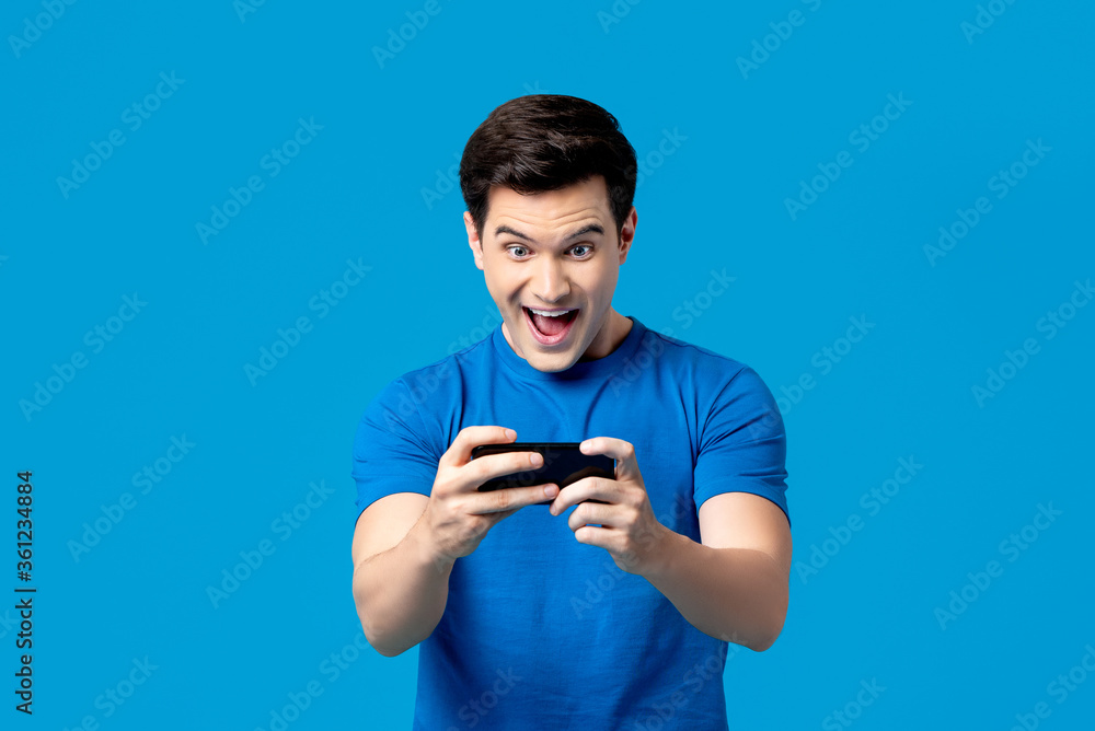 Excited handsome young male gamer playing online game on smartphone isolated on blue background Excited handsome young male gamer playing online game on smartphone isolated on blue background