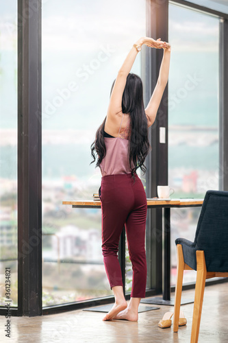 woman in casual clothes with arms raised standing and looking out of full-length window of apartment or hotel room.