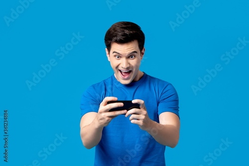 Excited handsome young male gamer playing online game on smartphone isolated on blue background