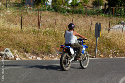 A spanish villager is riding a blue motorbike among farmlands, while carrying a barrel in front of him. He wears white underwear vest and a helmet. His arms are tanned due to direct sunlight.