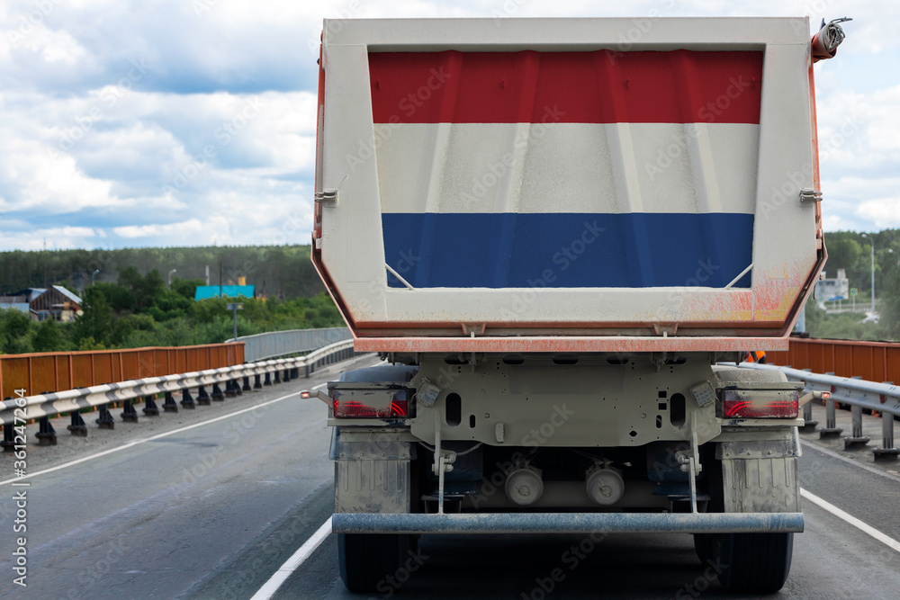 Big dirty truck with the national flag of Netherlands moving on the highway, against the background of the village and forest landscape. 