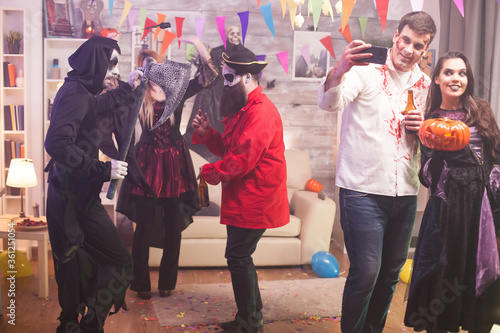 Beautiful witch holding a pumpkin at halloween party while zombie male is taking a selfie.