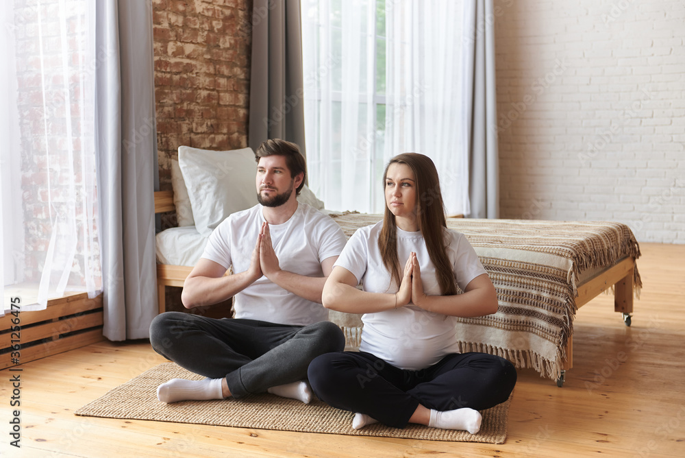 Young couple of beautiful pregnant woman and handsome husband practicing yoga and exercise, sitting with hands in Namaste, and meditating at home. Prenatal Yoga. Healthy maternity concept, fitness