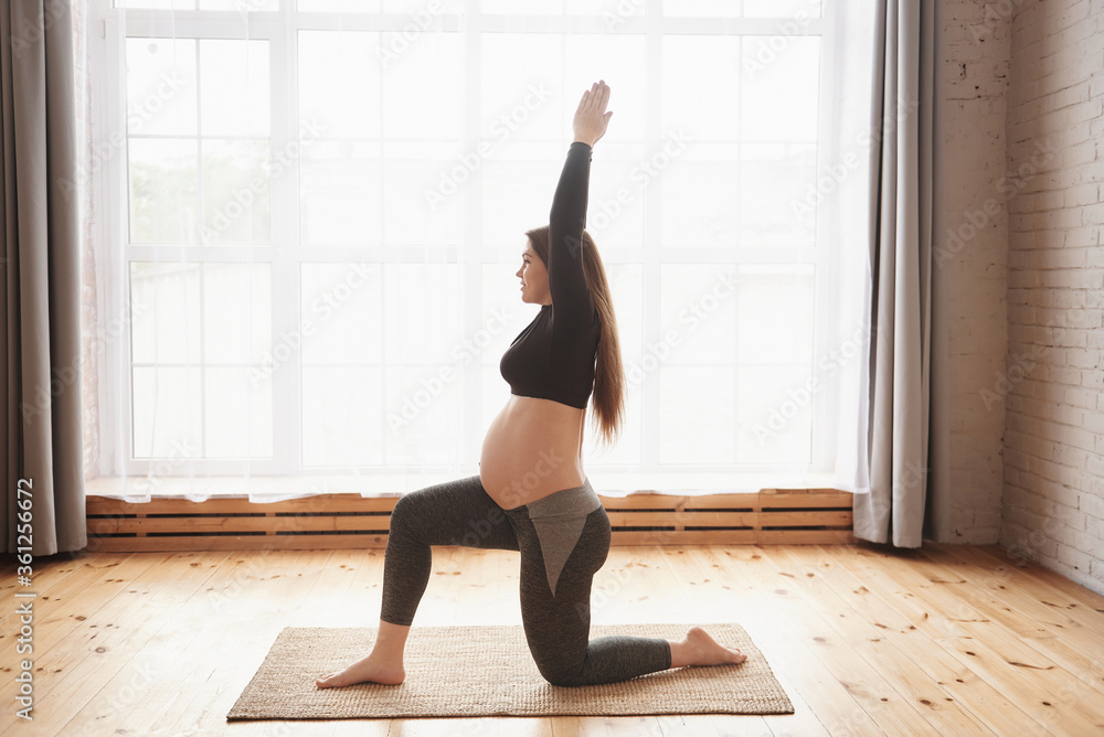 Young smiling pregnant woman, practicing yoga exercises at home or studio with big window. Prenatal variation of Warrior I posture. Pregnancy Yoga and Fitness concept. Full length, copy space.
