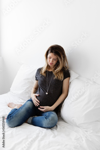 Pregnant woman sitting on white bed wearing bola necklace looking at her stomach photo