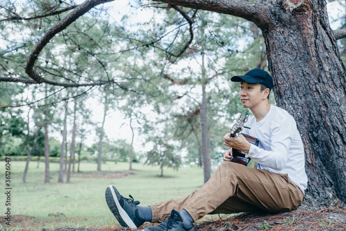 Asian man sitting and singing with guitar under pine tree.