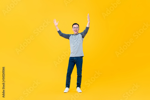 Full body portrait of cheerful young Asian man raising both arms in the air isolated on yellow background