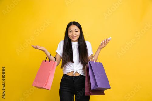 Showing, uncertainty. Young asian woman with colorful shopping packages on yellow studio background. Stylish, trendy. Beautiful brunette. Human emotions, facial expression, sales, ad, shopping concept
