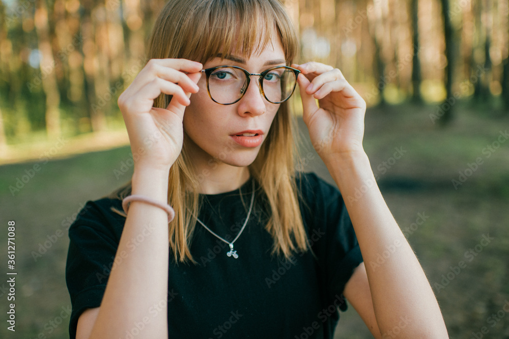 Portrait of beautiful young girl with short fair hair, blue eyes, black glasees in black jacket poses for the camera in the park Portrait of beautiful young girl with short fair hair, blue eyes, black glasees in black jacket poses for the camera in the park