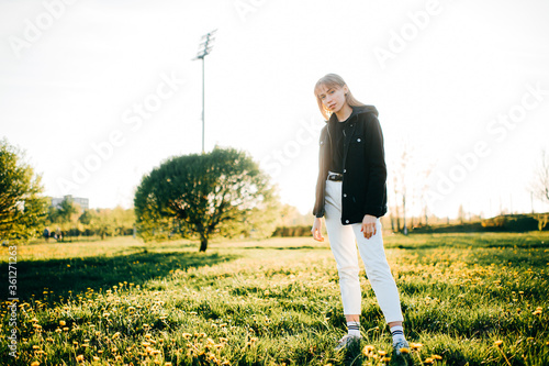 Beautiful young woman with short fair hair and blue eyes in black jacket goes for a walk to the summer park in sunny weather