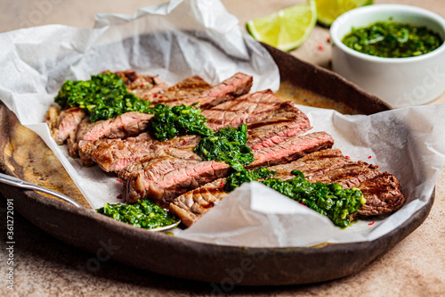 Sliced grilled beef steak with chimichurri sauce on dark dish, dark background.