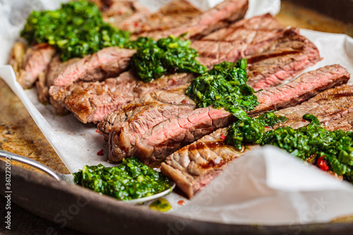 Sliced grilled beef steak with chimichurri sauce on dark dish, dark background, close-up.