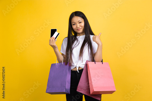 Online purchase. Young asian woman with colorful shopping packages on yellow studio background. Stylish, trendy. Beautiful brunette. Human emotions, facial expression, sales, ad, shopping concept.