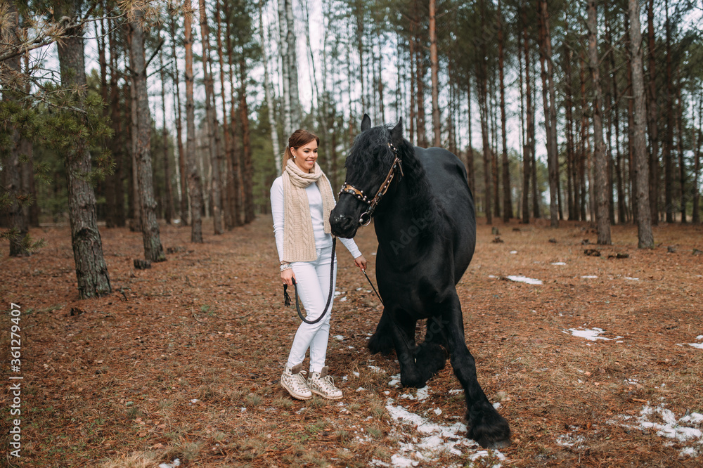 Magnetic young female in grey fur coat and white trousers stands with a big black horse outside Magnetic young female in grey fur coat and white trousers stands with a big black horse outside