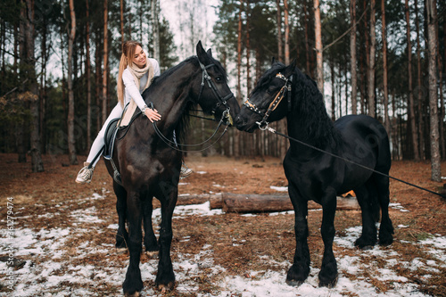 Handsome caucasian woman with fair hair in warm clothes goes for a walk with two black horses in winter forest. © benevolente