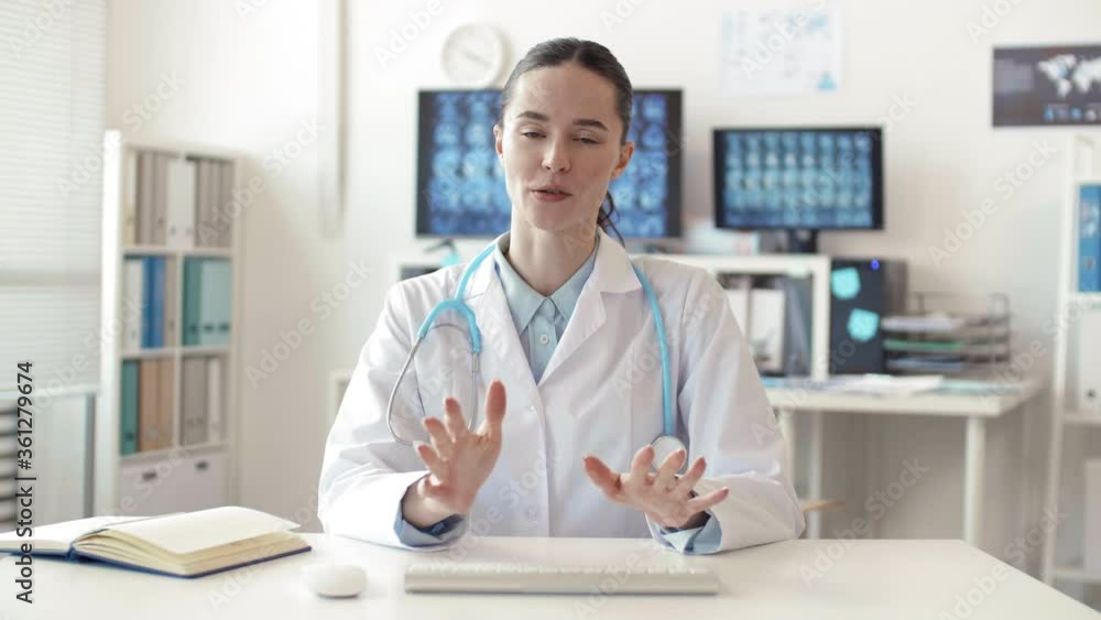 Medium shot portrait of young Caucasian woman wearing medical overall and stethoscope sitting in front of computer, communicating with online patient and typing on wireless keyboard Medium shot portrait of young Caucasian woman wearing medical overall and stethoscope sitting in front of computer, communicating with online patient and typing on wireless keyboard