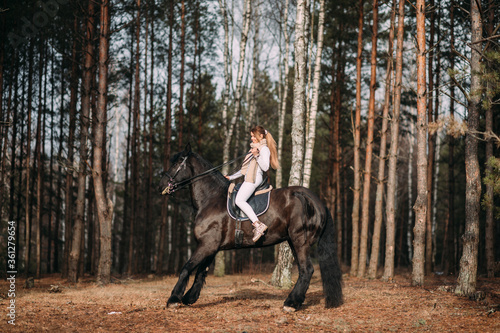 Bizzare young female in grey fur coat and white trousers training for rides on a big black horse outside.