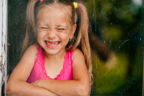 Little cute girl with blond hair and pink dress smiles behind the window © benevolente