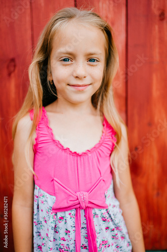 Little cute girl with blond hair and pink dress makes faces against the wooden wall