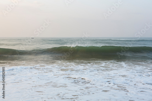 Fototapeta Naklejka Na Ścianę i Meble -  natural copy space shot of a wave crushing on the smooth yellow sand beach surface, forming white foam, with the blue sky on the horizon. Early morning sunrise at Pitiwella beach, Sri Lanka