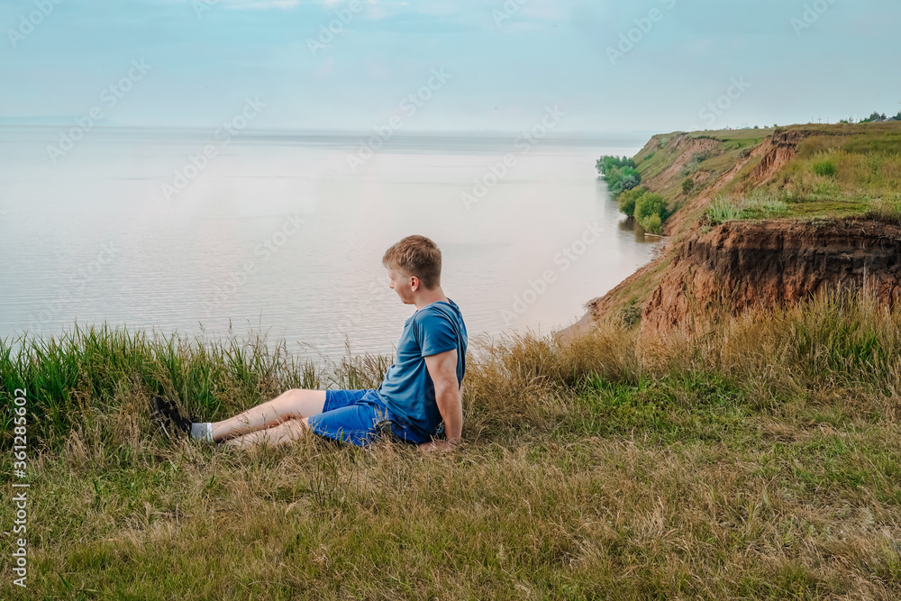 A blond man sits on the grass in front of a rock from the sea