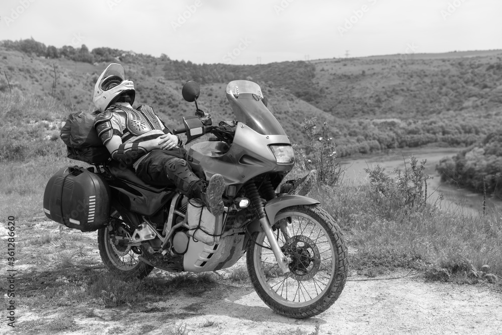 Naklejka premium Motorcycle driver resting after a long and difficult road, lies on a motobike, wearing a turtle jacket, body armor. on top. View of the forest and river. Black and white