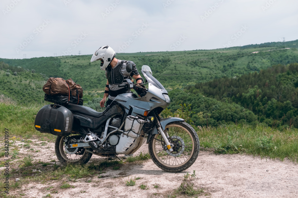 The guy is a motorcycle driver wearing a turtle jacket, body armor and a white helmet. Stands on top. View of the forest and sky. The concept of freedom of new impressions and tourism. Extreme hobby
