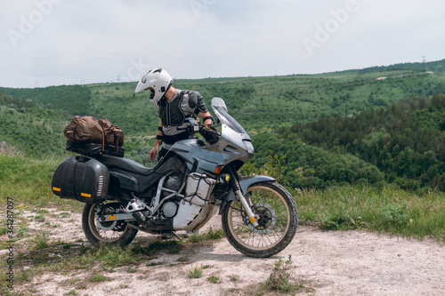 Fototapeta Naklejka Na Ścianę i Meble -  The guy is a motorcycle driver wearing a turtle jacket, body armor and a white helmet. Stands on top. View of the forest and sky. The concept of freedom of new impressions and tourism. Extreme hobby