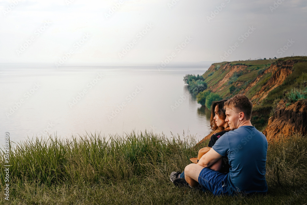Fototapeta premium A back view of young adult lovers sitting on the edge of a cliff and watching the sea on a rainy and gloomy day. the concept of travel.